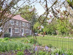 Un bâtiment en pierre, surmonté d’un toit recouvert de tuiles rouges, entouré d’un jardin bien entretenu où poussent différentes espèces de plantes et délimité par une clôture en métal.