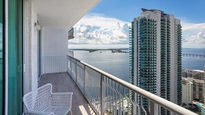 Un balcon avec une chaise blanche, donnant sur un immeuble de grande hauteur et sur un pont enjambant une étendue d’eau.