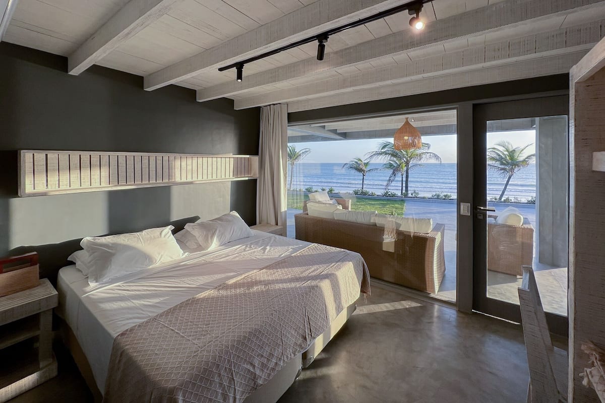 A modern bedroom with a large bed, a headboard with a built-in shelf, a view of the ocean, and a balcony with outdoor seating.