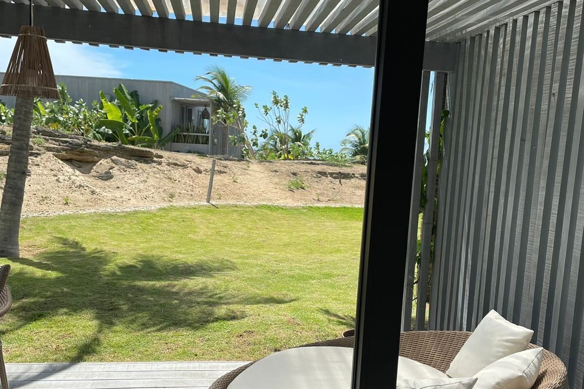 A wicker lounge chair with cushions on a wooden deck, a covered patio area, and a view of a grassy yard with a building in the background.