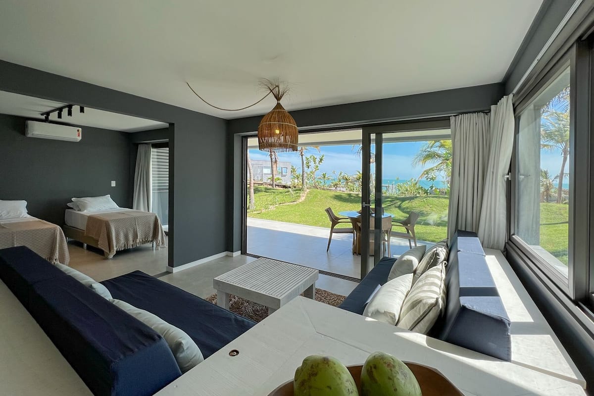 A modern hotel room with a large sliding glass door leading to a balcony, a bed, a sofa, and a dining table with two fruits on it.