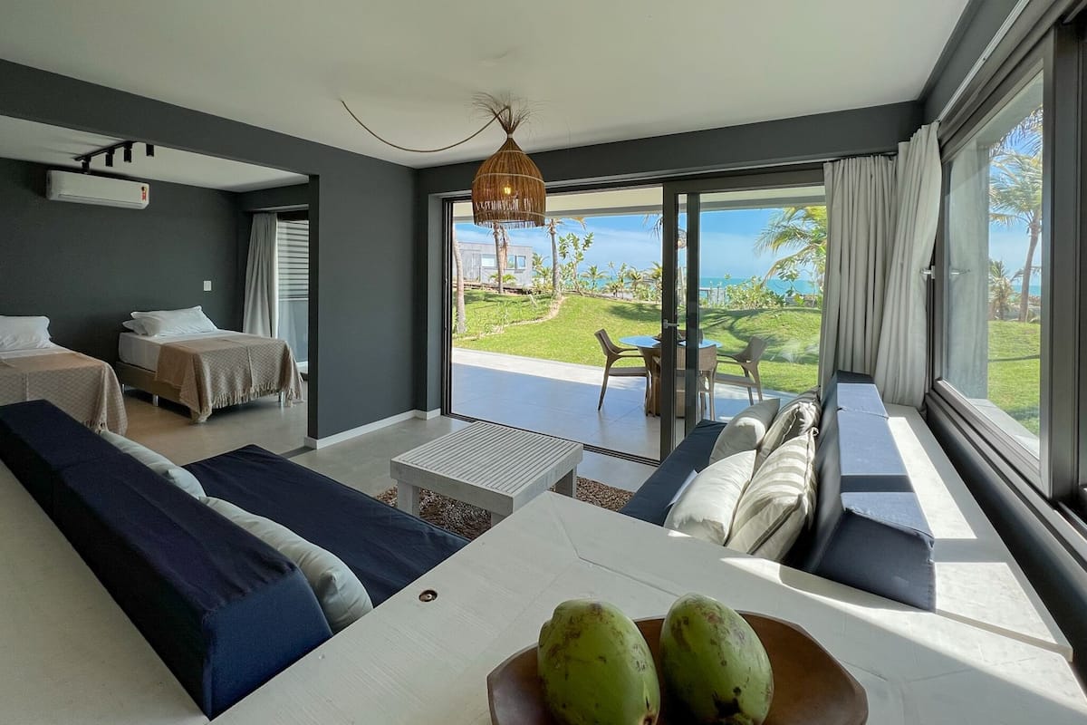 A modern hotel room with a large sliding glass door leading to a balcony with a view of the ocean and palm trees. There are two beds, a sofa, and a dining table with a bowl of fruit.