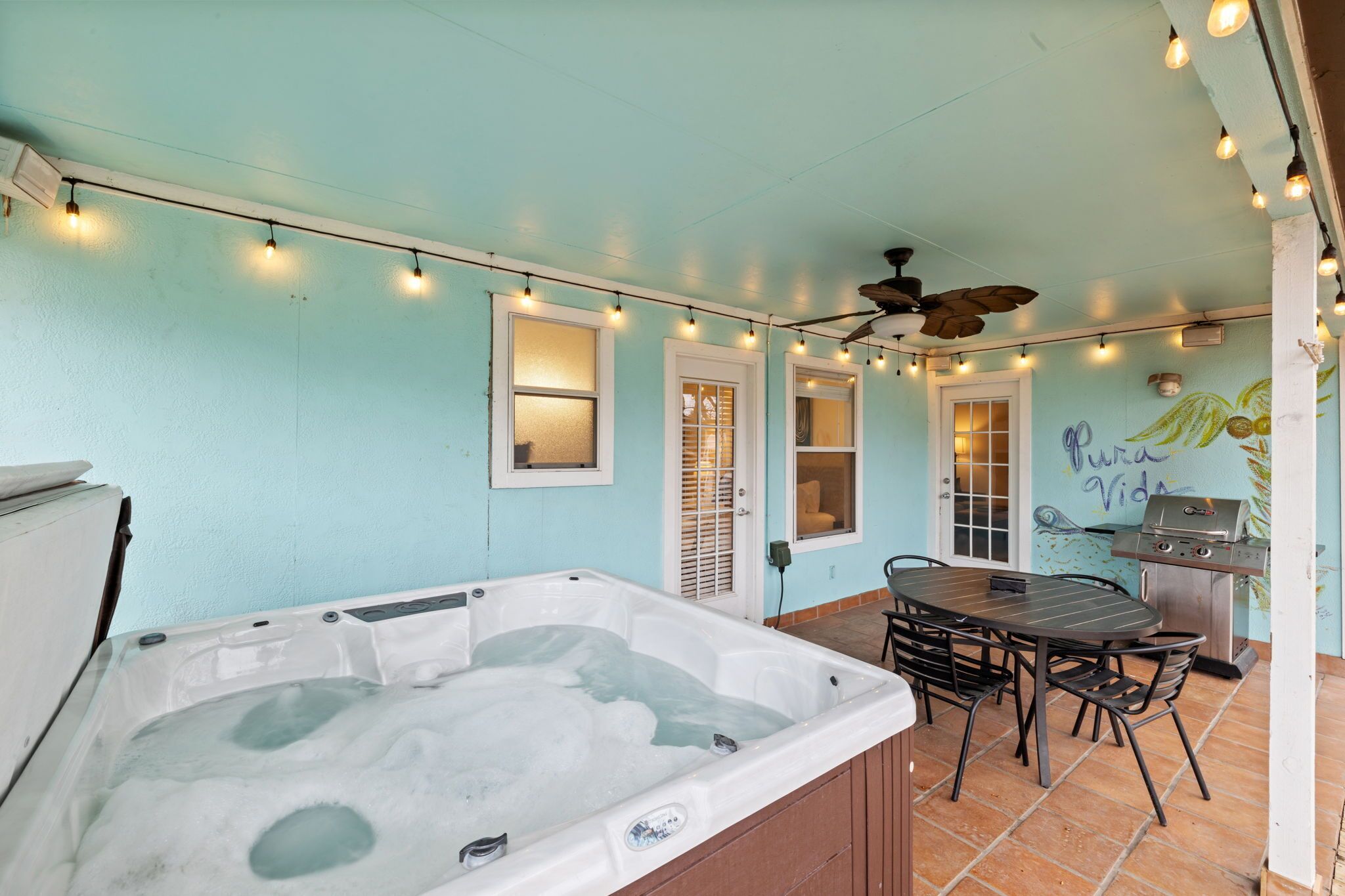 A covered patio with a hot tub, a dining table, chairs, string lights, and a grill.