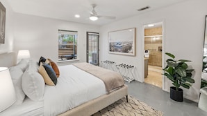 Quarto moderno com cama, luminárias de cabeceira, ventoinha no teto, janela com persianas, porta para um banheiro e um vaso de planta.