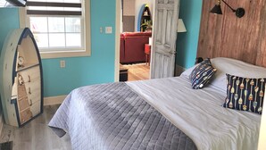 Quarto com cama, ventoinha no teto, janela com persianas, guarda-roupa de madeira e porta que dá para outro cômodo.