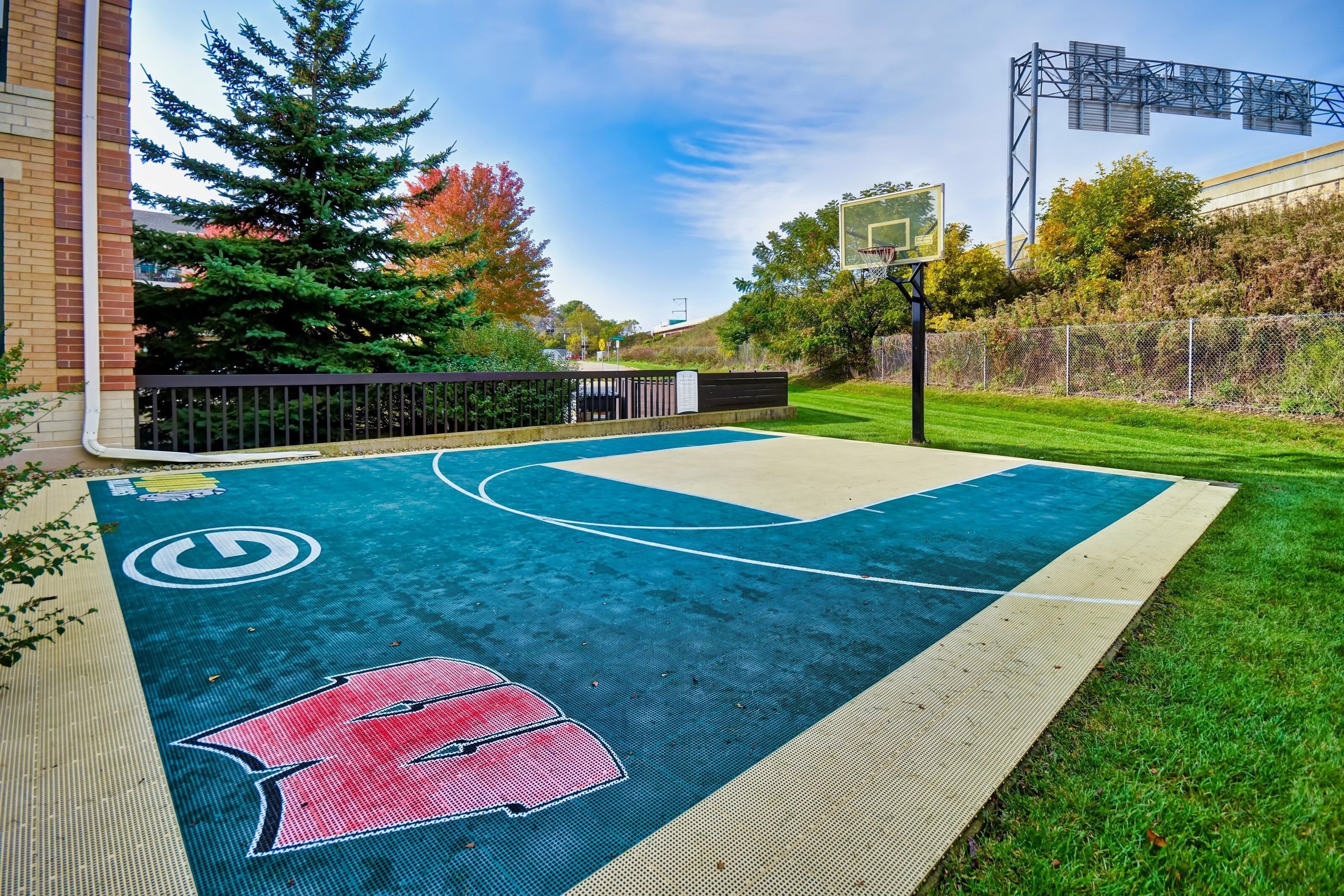 An outdoor basketball court with a red logo, surrounded by a fence, trees, and a building in the background.