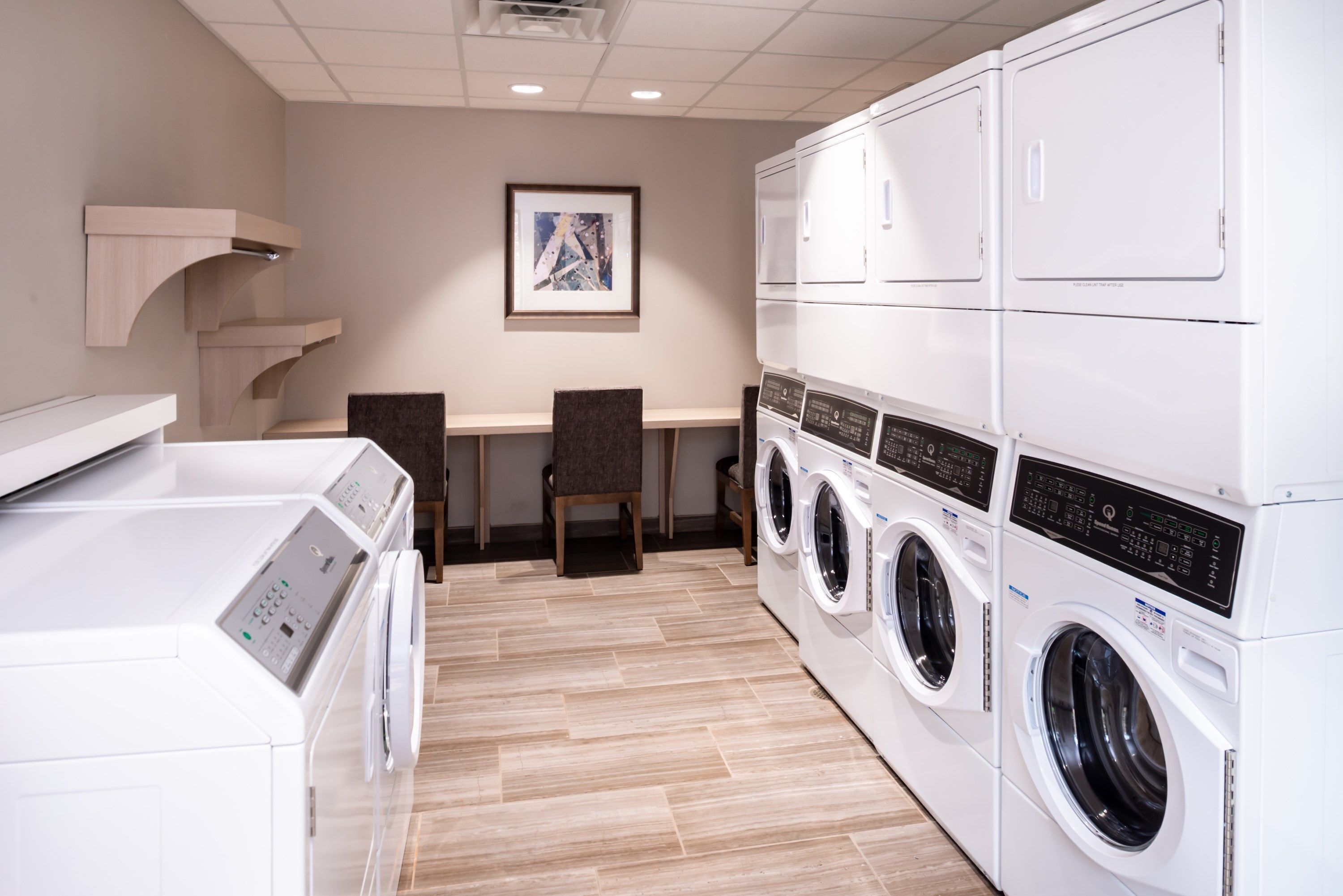 A laundry room with stacked washing machines and dryers, a wooden chair, and a framed picture on the wall.