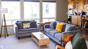 Ein modernes Wohnzimmer mit zwei grauen Sofas, einem Holztisch und einem kleinen Holzschemel. Ein Küchenbereich mit Holzschränken und einem Barhocker vorhanden.