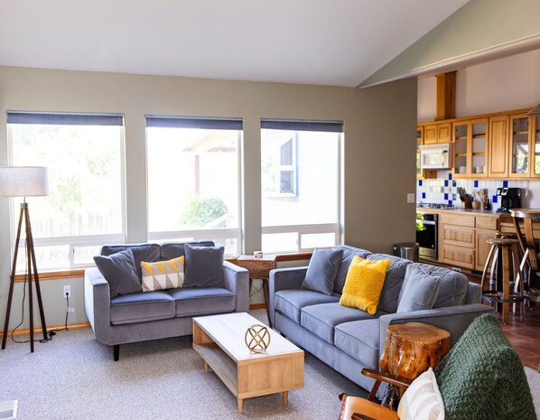 A modern living room with two grey sofas, a wooden coffee table, and a small wooden stool. There is a kitchen area with wooden cabinets and a bar stool.