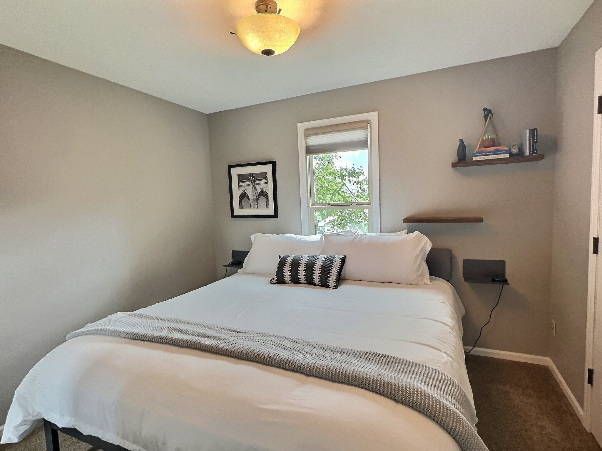 A bedroom with a large bed, a window, a framed picture, and a shelf with books.
