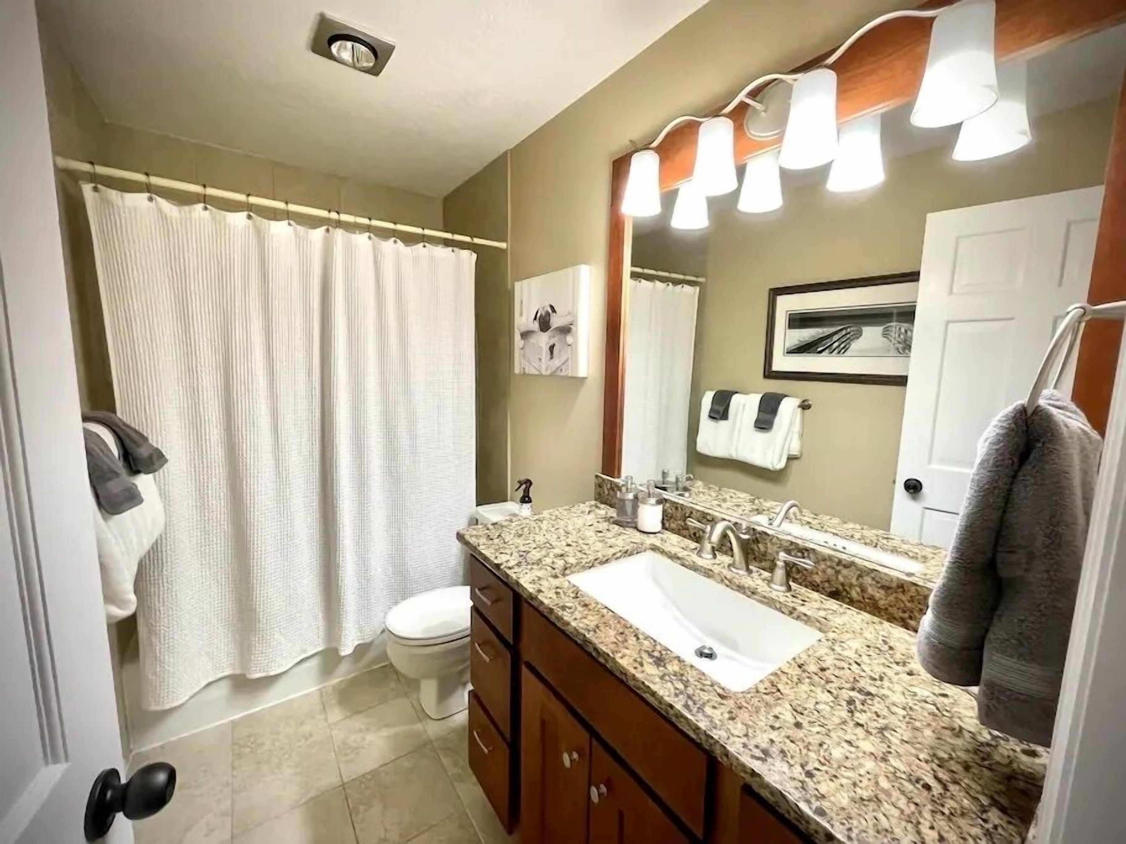 A bathroom with a shower curtain, toilet, sink, vanity, and a mirror with lights.