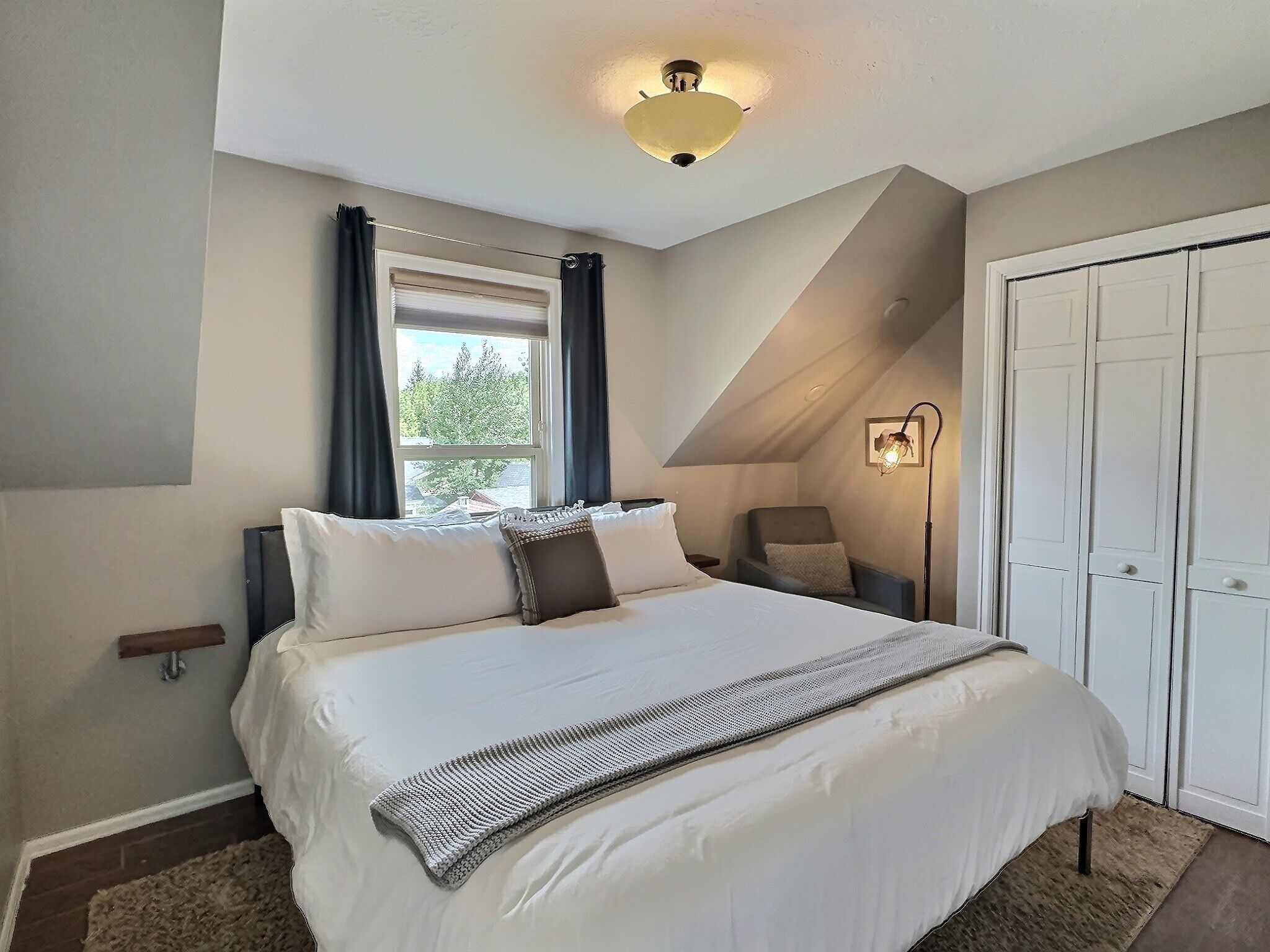 A bedroom with a large bed, a window with curtains, a chair, a bedside table, and a wardrobe.