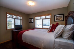 Un dormitorio con una cama grande, dos ventanas con persianas, un televisor sobre un mueble bajo y un cuadro enmarcado en la pared.
