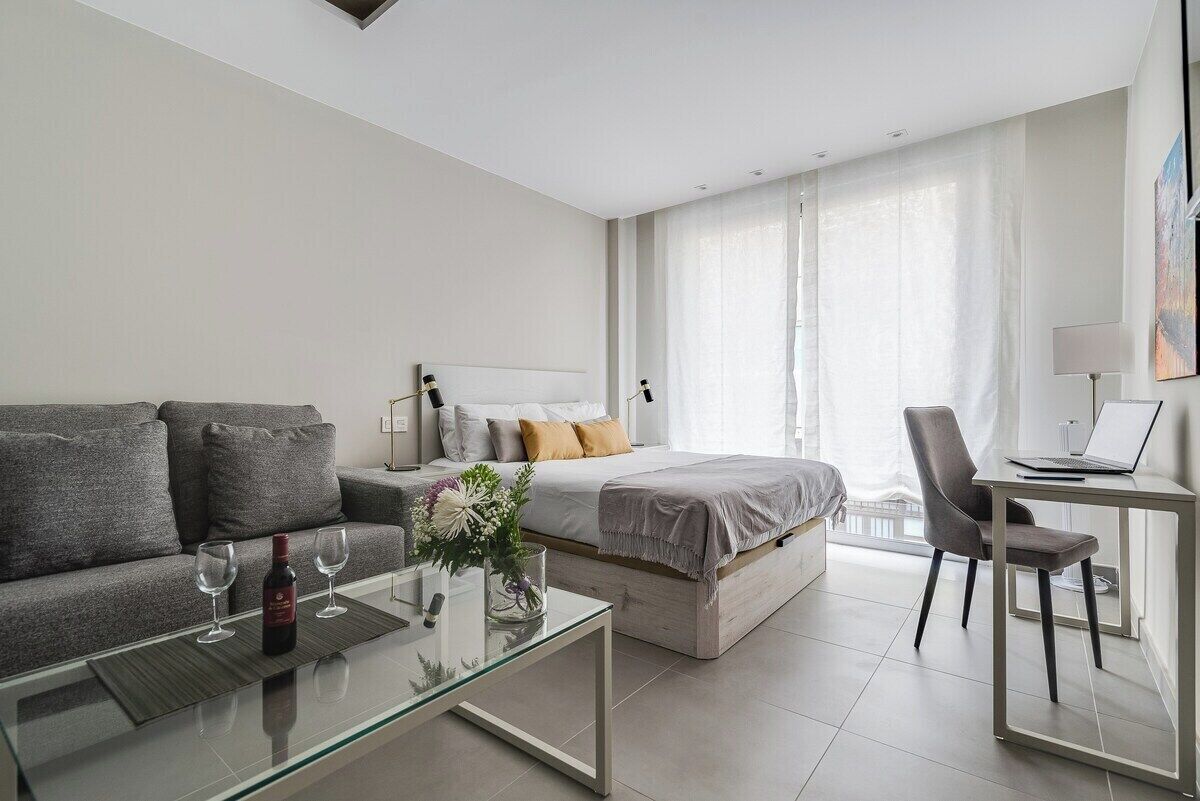A modern hotel room with a sofa, a bed, a glass coffee table, a desk with a laptop, and a large window with curtains.