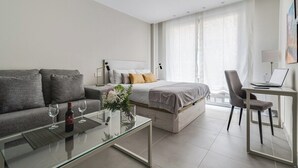 Une chambre d’hôtel moderne équipée d’un canapé, d’un lit, d’une table basse en verre, d’un bureau avec un ordinateur portable et d’une grande fenêtre avec des rideaux.
