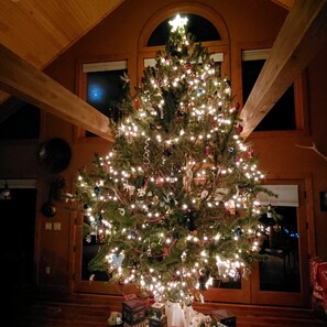 Un árbol de Navidad decorado con luces y adornos, situado en una habitación con vigas de madera y un ventanal.