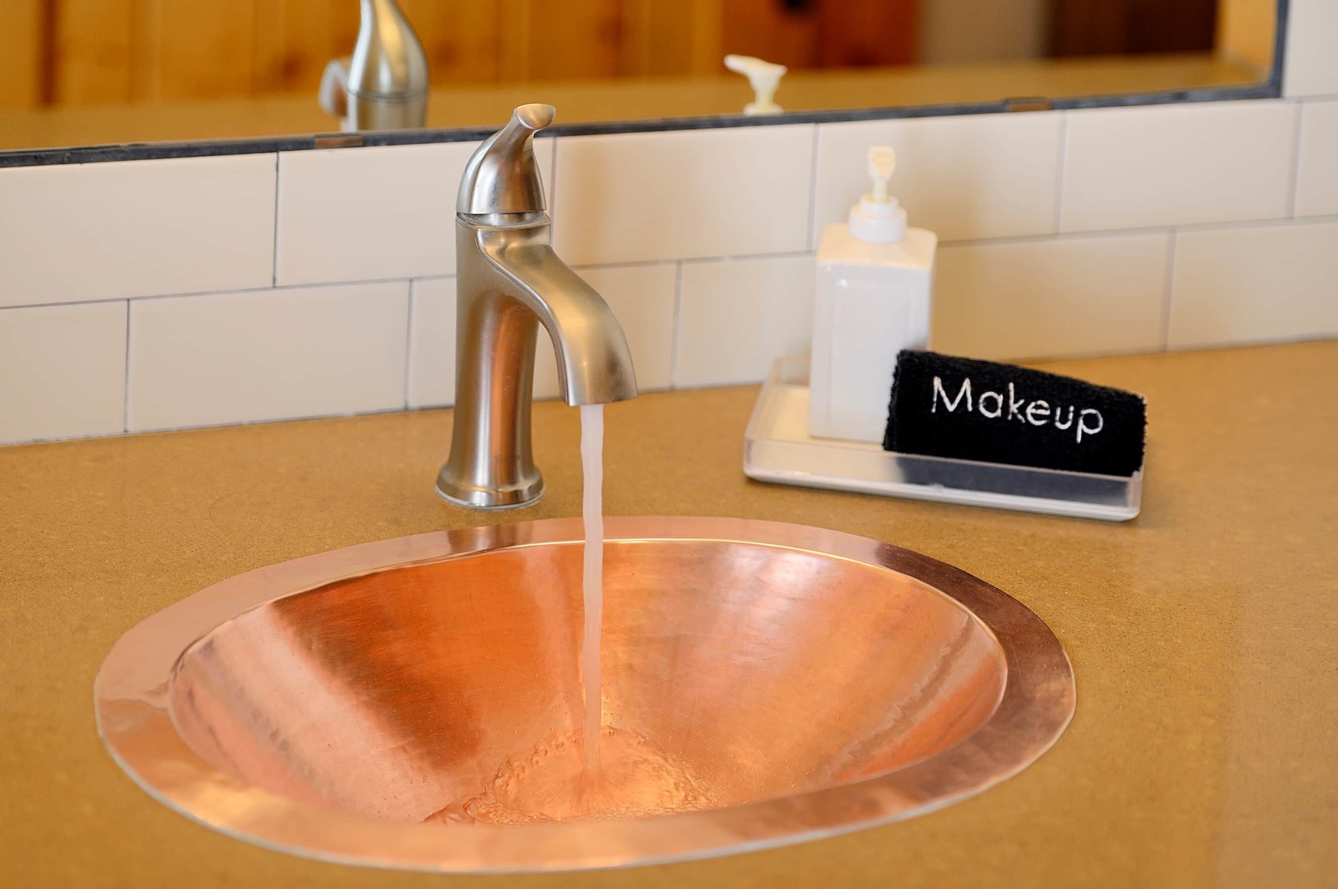 A copper sink with running water, a makeup station, and a mirror in a bathroom.