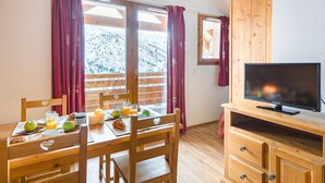 A wooden dining table with breakfast items, a TV on a wooden stand, and a view of a snowy mountain through large windows. - Près des pistes | séjour avec accès piscine + spa! (Valmeinier)