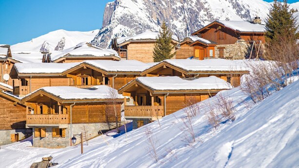 A row of wooden chalets with snow-covered roofs, set against a backdrop of snow-capped mountains. - Près des pistes | séjour avec accès piscine + spa! (Valmeinier)