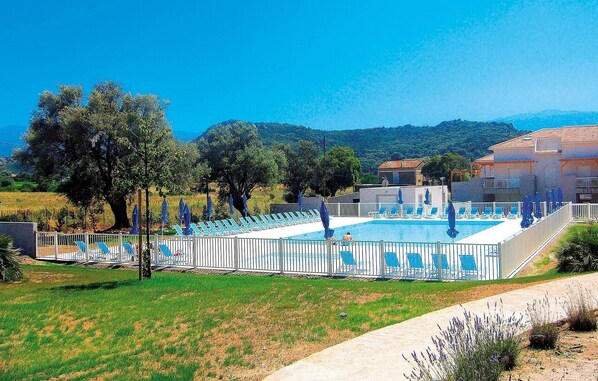 Una piscina con sillas de descanso, rodeada por una cerca blanca, con edificios y árboles al fondo.