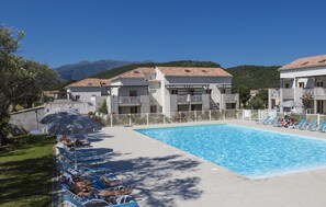 Una piscina con sillones de descanso, un área techada con sombrillas y edificios con balcones.