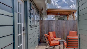 A covered patio with two orange chairs and a table, a wooden fence, and a house with blue siding and a white door. - Quiet, Cozy Townhouse w/ all new appliances and furniture. (Shreveport)