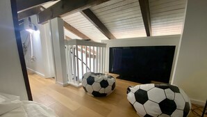 Una habitación con techo alto, vigas de madera, una escalera, un televisor grande y dos pufs con forma de balones de fútbol.
