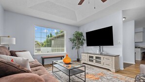 Un salon avec un canapé marron, une table basse en verre, un téléviseur, une plante et, au fond, un coin cuisine.