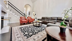 A spacious living room with a large sofa, two armchairs, a coffee table, and a staircase with a chandelier.