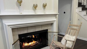 A fireplace with a fire, a wooden rocking chair with a fluffy cushion, and a staircase in the background.