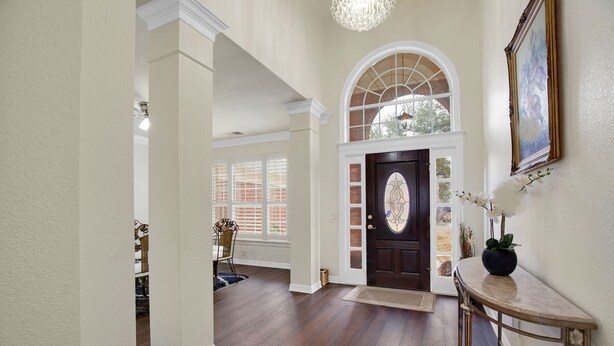 A spacious foyer with a wooden floor, a dark wooden door with a decorative glass panel, and a chandelier.
