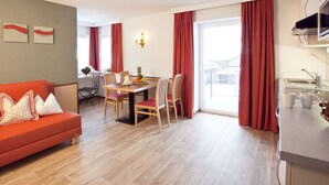 Ein modernes Wohnzimmer mit einer Couch, einem Esstisch und Stühlen. Es gibt eine Küchenzeile mit Spüle und Schränken. Der Raum verfügt über große Fenster mit roten Vorhängen.