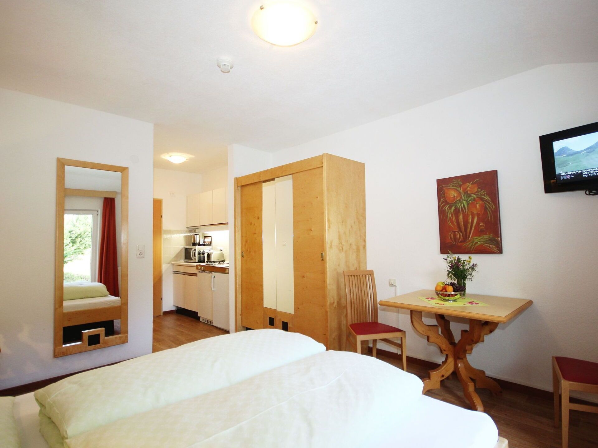 Hotelkamer met een bed, een houten tafel met twee stoelen, een televisie, een schilderij en een keukenruimte zichtbaar door een geopende deur.