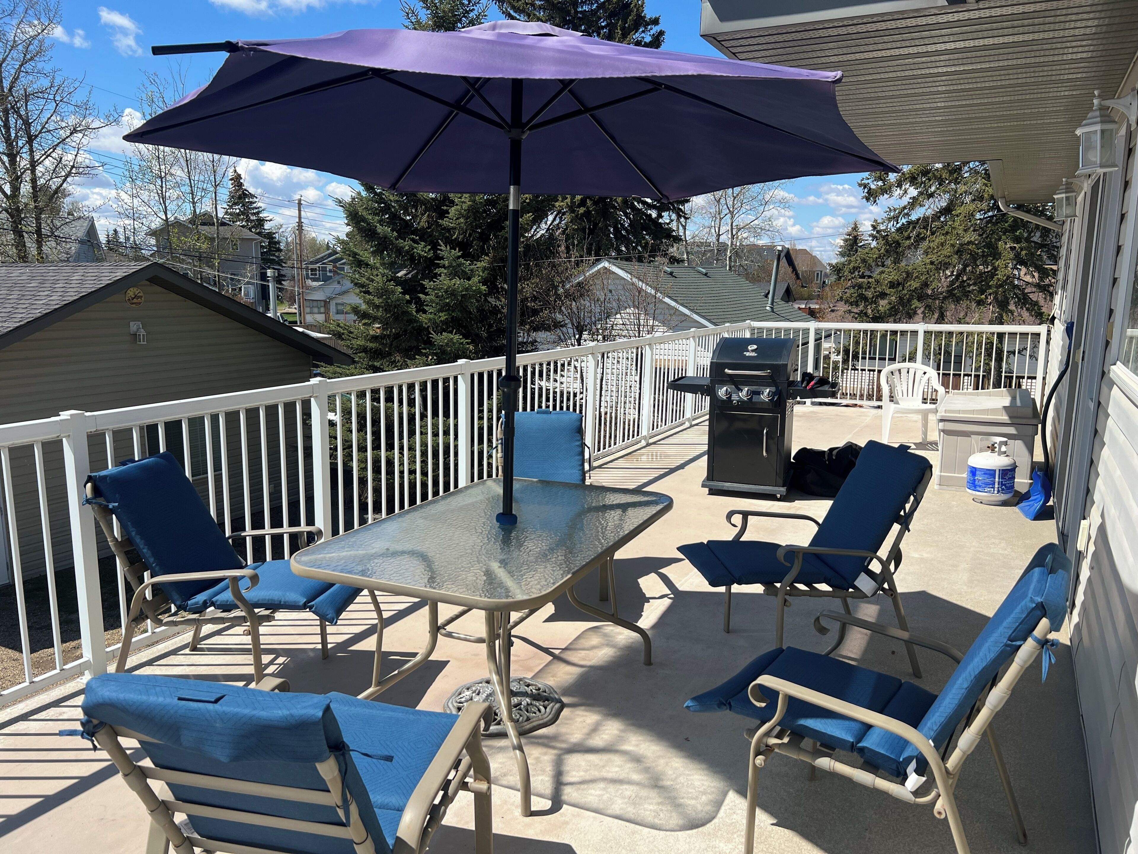 An outdoor patio with a table and chairs, a barbecue grill, and a white railing.