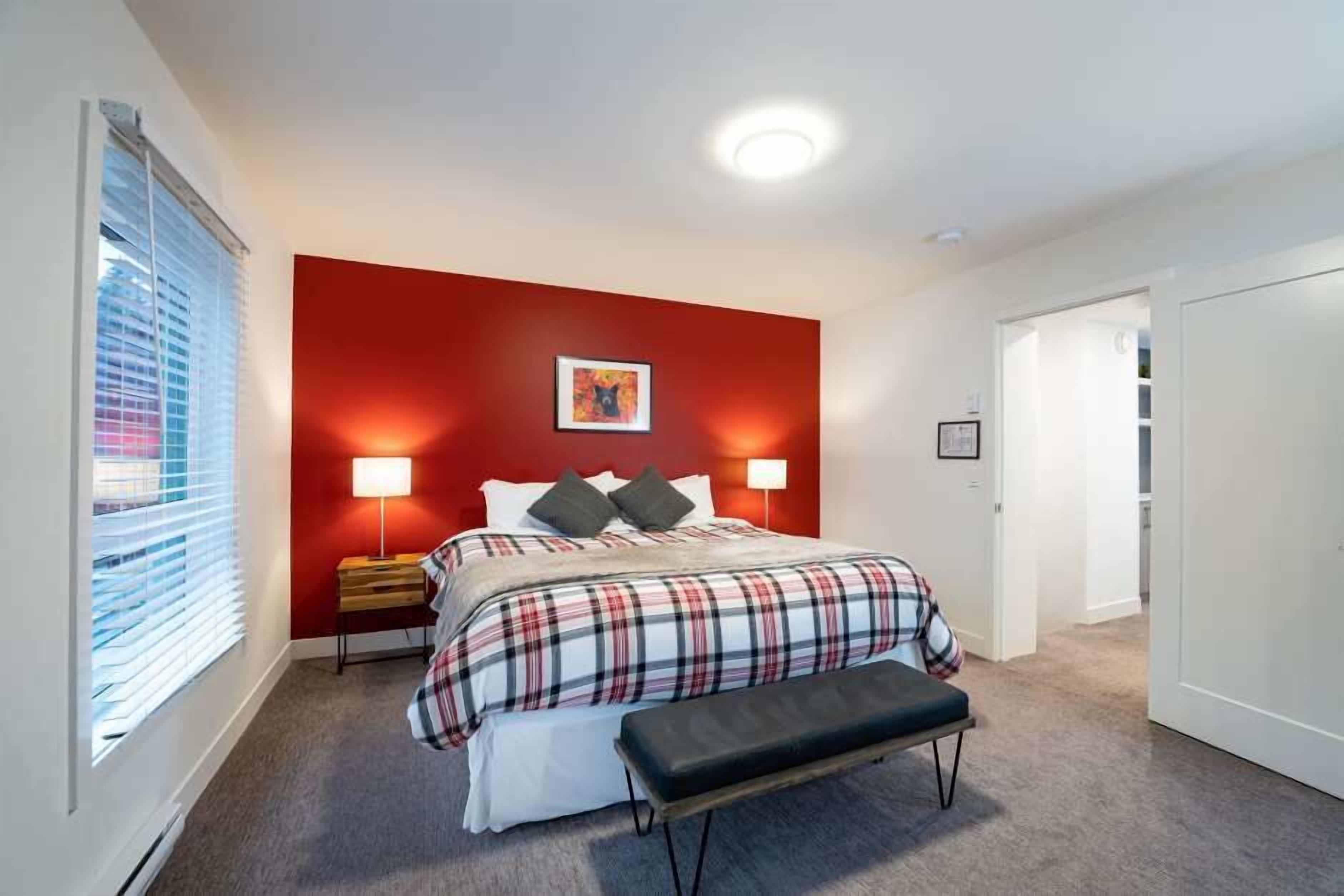 Een slaapkamer met een rode accentmuur, een bed met een geruit beddegoed, twee bedlampen, een bankje en een ingelijste foto aan de muur.