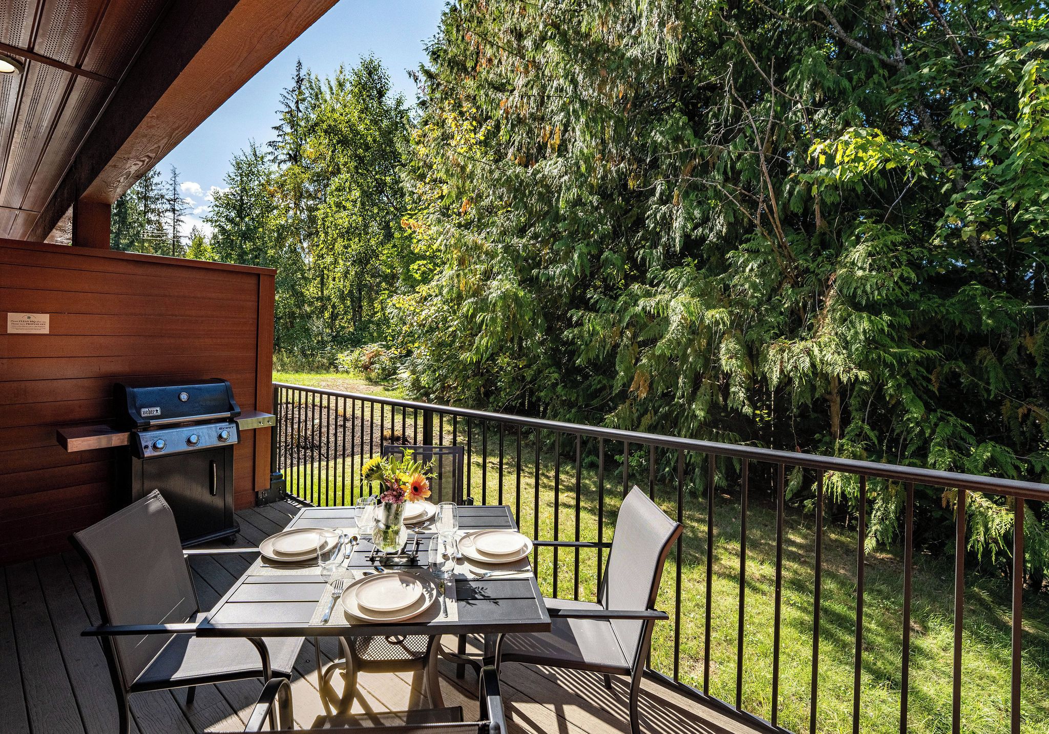 An outdoor deck with a grill, dining table, chairs, and a view of a forested area.