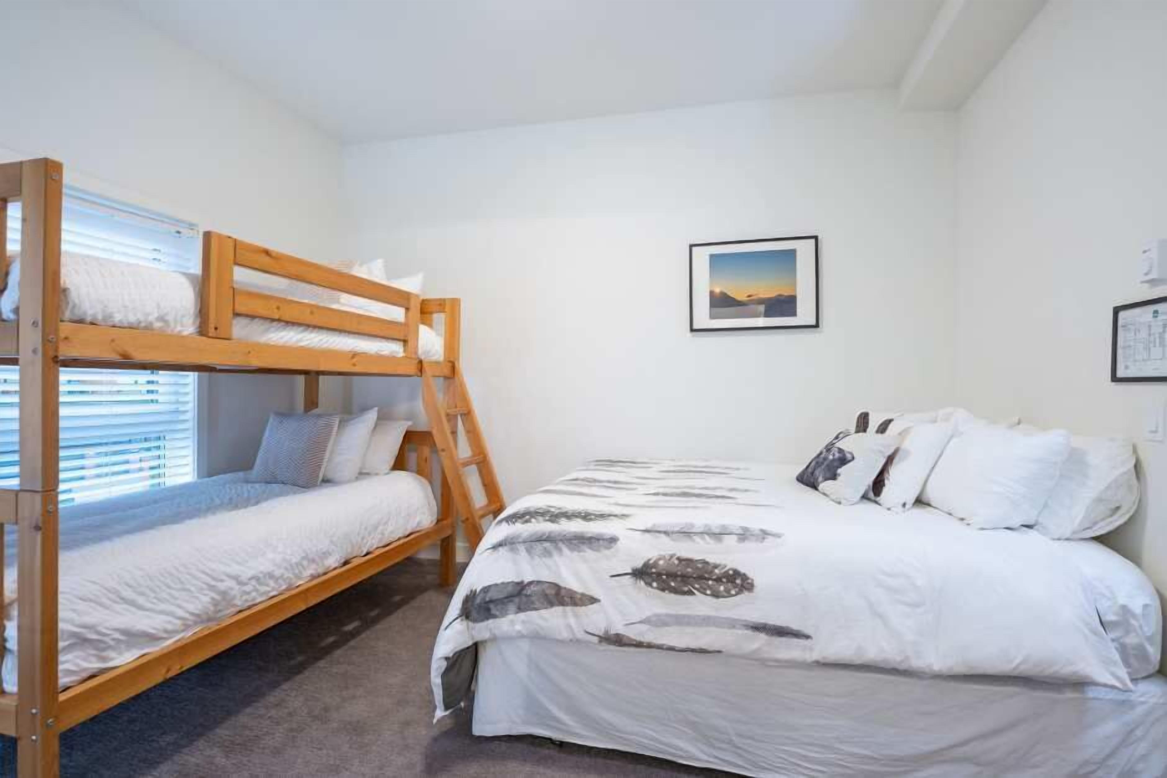 A bedroom with a bunk bed and a single bed, both with white bedding. There is a framed picture on the wall and a ladder beside the bunk bed.