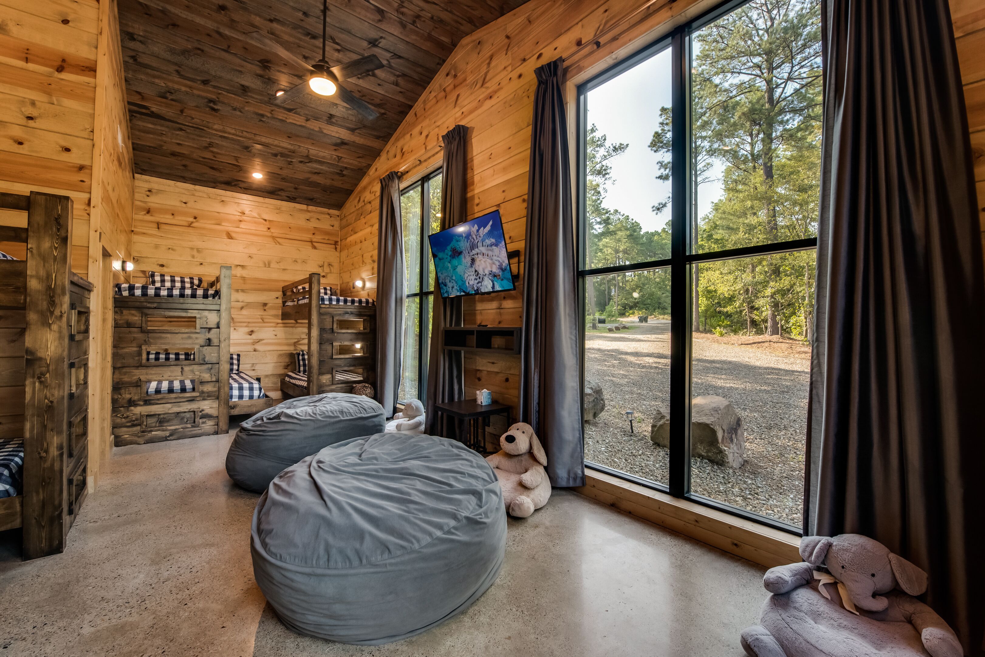 A cozy cabin with wooden walls, large windows, and a TV on the wall. There are bean bags and a stuffed animal on the floor.