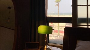 Habitación de hotel con cama, mesita de noche con lámpara, ventana con vista a la playa y una palmera, y una tabla de surf en la pared.
