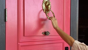Una mano alcanza un llamador dorado en una puerta rosa, con una cerradura y un pequeño pomo rectangular debajo.