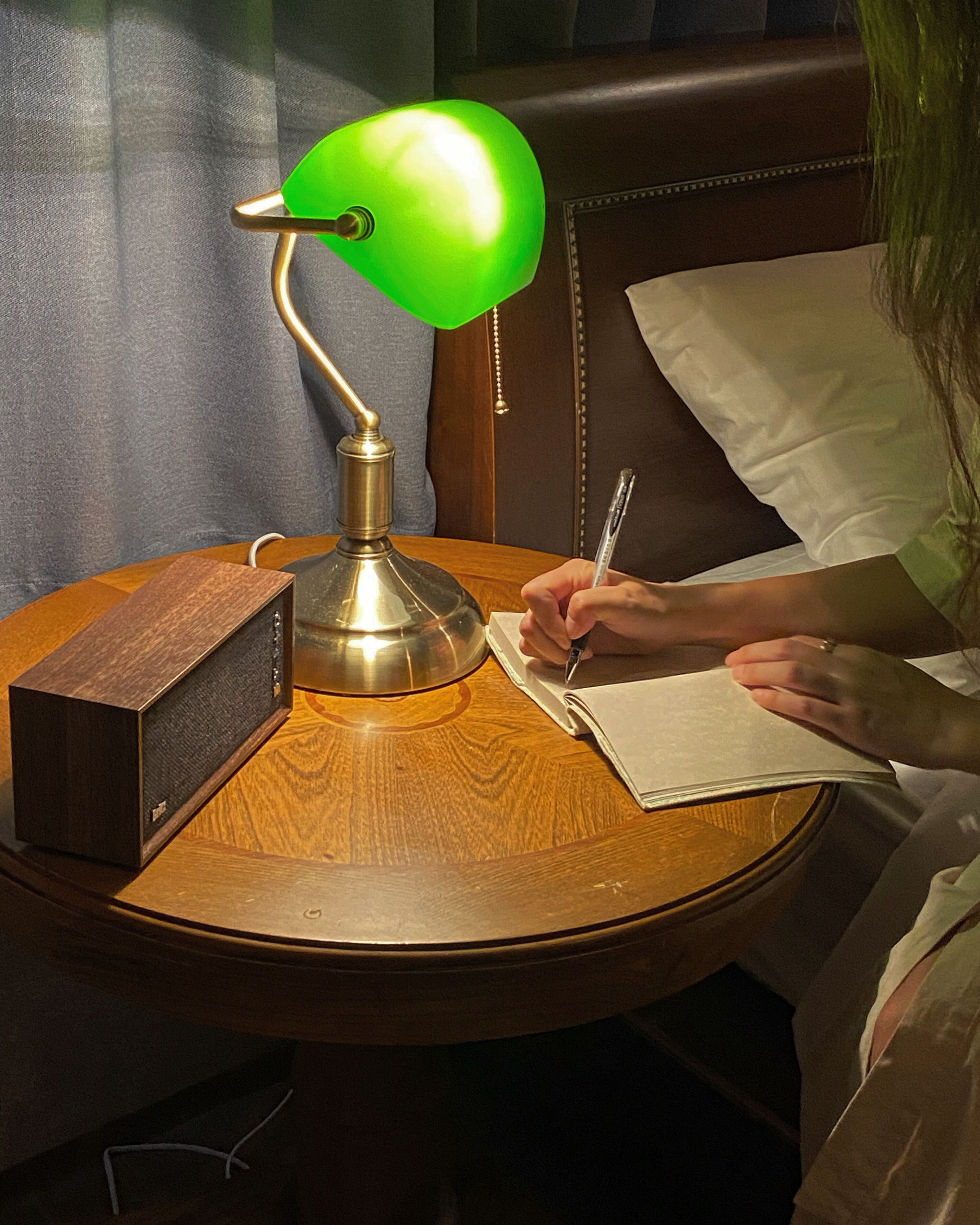 A round wooden table with a green desk lamp, a radio, and a person writing in a notebook.