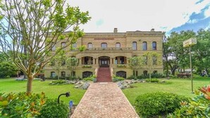 Un edificio histórico con una entrada central, un sendero de ladrillo que lo conduce y jardines bien cuidados.