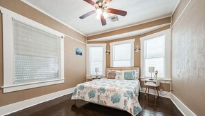Un dormitorio con cama, ventilador de techo y dos ventanas con persianas.
