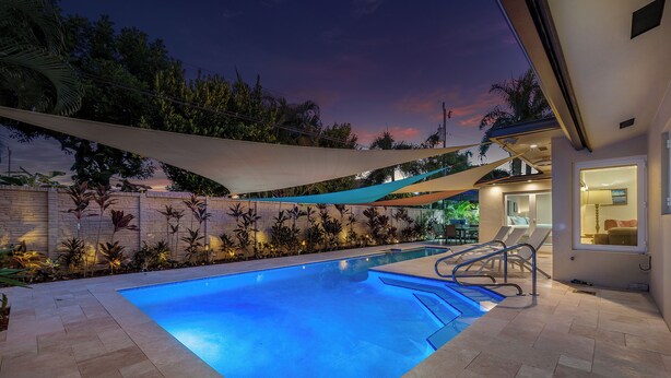 Ein Pool mit klarem, blauem Wasser, umgeben von einer gefliesten Terrasse und einem schattigen Bereich mit einer weißen Wand und Pflanzen.