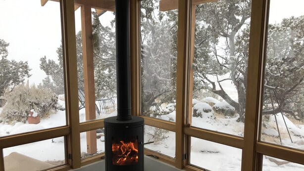 A wood-burning stove in a room with large windows, a wooden ceiling, and a view of a snowy landscape.