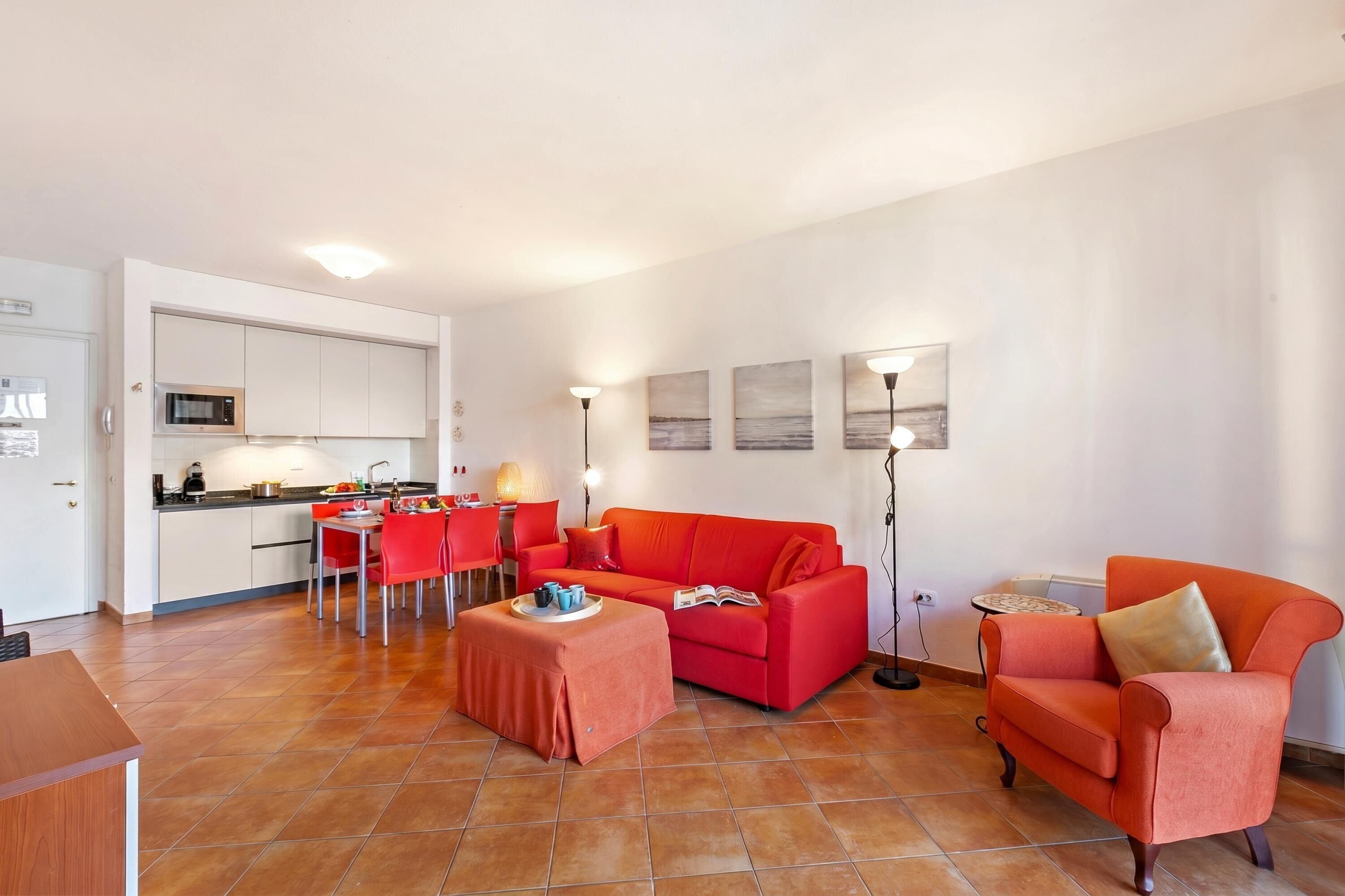 Ein Wohnzimmer mit einem roten Sofa, einem Couchtisch, einem Essbereich mit roten Stühlen und einer Küche mit weißen Schränken und einer Mikrowelle.