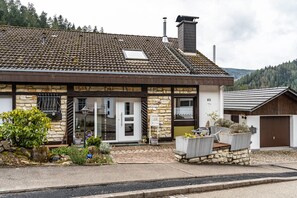 Un bâtiment en pierre avec un toit en tuiles, une porte d’entrée et un petit jardin agrémenté de plantes et d’un banc.