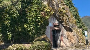 Un edificio con fachada de piedra, un pequeño arco de entrada y una ventana con contraventanas verdes.