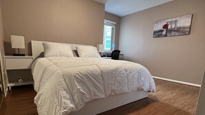 Et soveværelse med en stor seng, et sengebord med lampe, en stol, et vindue og et billede på væggen.