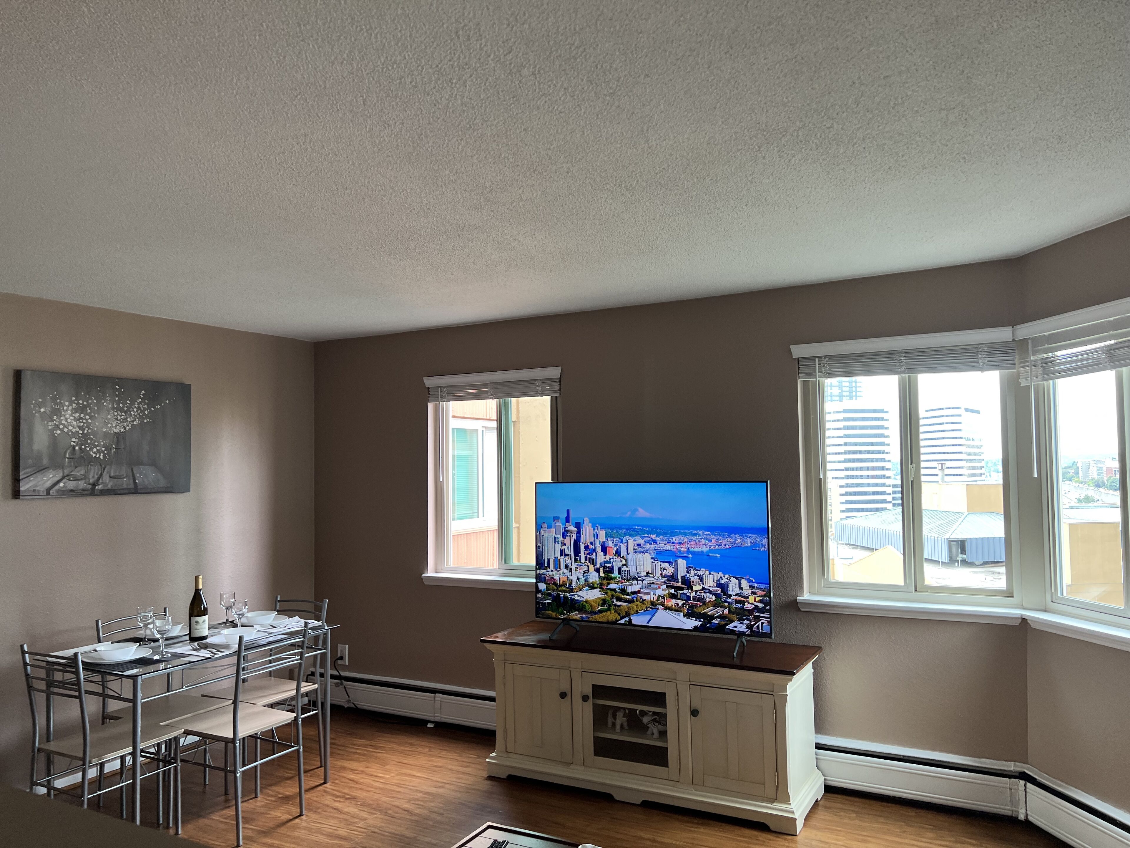 Ein Wohnzimmer mit Essbereich, einem Flachbildfernseher und Blick auf eine Stadtkulisse durch große Fenster.